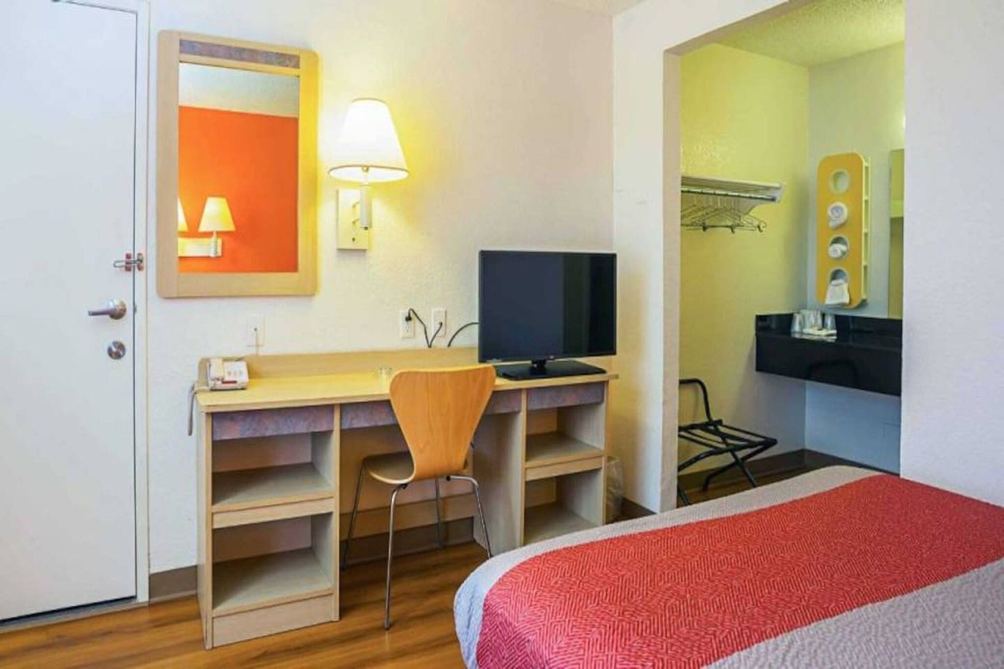 Habitación de hotel con escritorio, silla, televisión y cama con cubrecama rojo y gris.