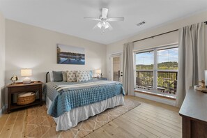 A bedroom with a bed, nightstands, a ceiling fan, a window with blinds, and a view of a dock and trees. - Lakeside Leisure Lake Views (Branson)