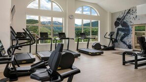 Une salle de sport équipée de tapis de course, de vélos elliptiques et d’un vélo d’appartement, de grandes fenêtres offrant une vue sur les montagnes et d’un mur sur lequel est projetée une œuvre d’art numérique.