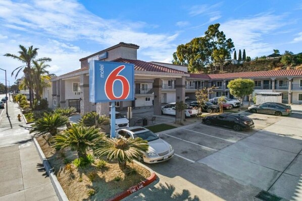 Un motel avec un grand panneau indiquant le chiffre « 6 », un parking avec plusieurs voitures et un bâtiment doté d’un toit recouvert de tuiles rouges.
