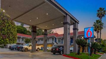 A motel with a covered parking area, multiple cars parked, and a palm tree in the foreground.