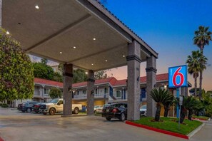 Un motel con área de estacionamiento techada, varios coches estacionados y una palmera en primer plano.