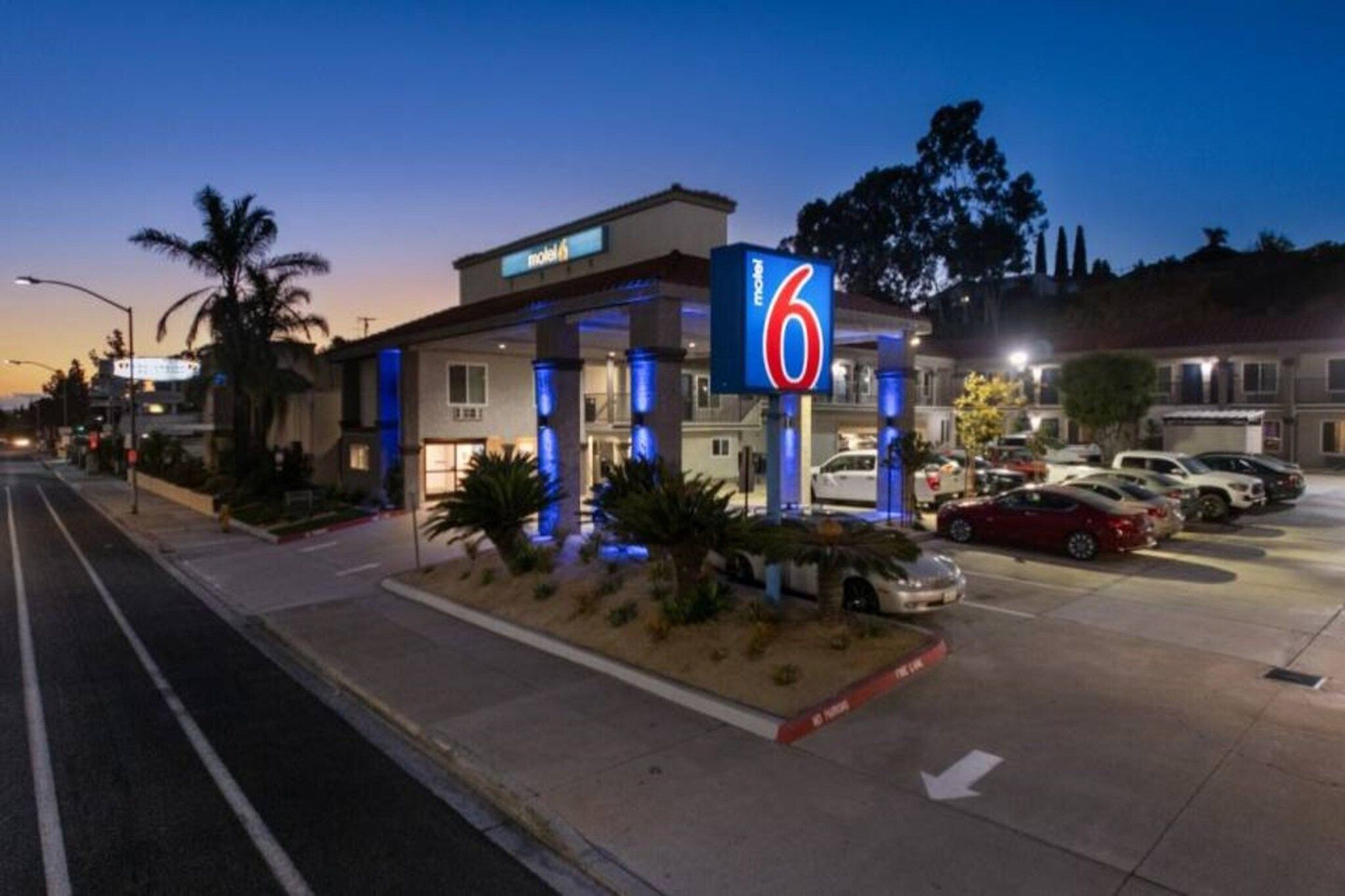 Un motel con un cartel llamativo, un estacionamiento con varios coches y una calle con palmeras.