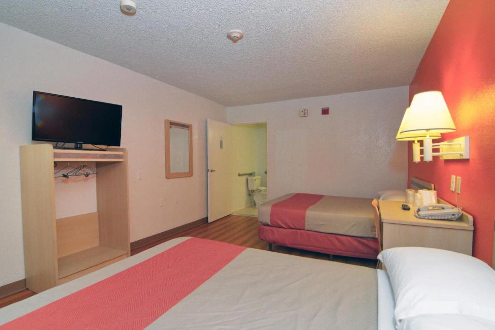 A hotel room with a bed, a television, a bedside table with a lamp, and a bathroom visible through an open door.