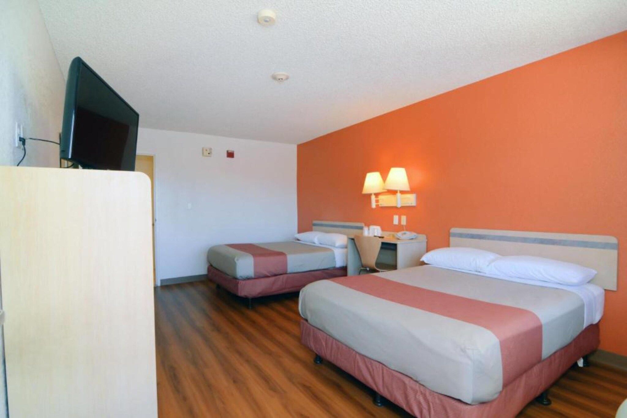 A hotel room with two beds, a television, and a wall-mounted lamp.