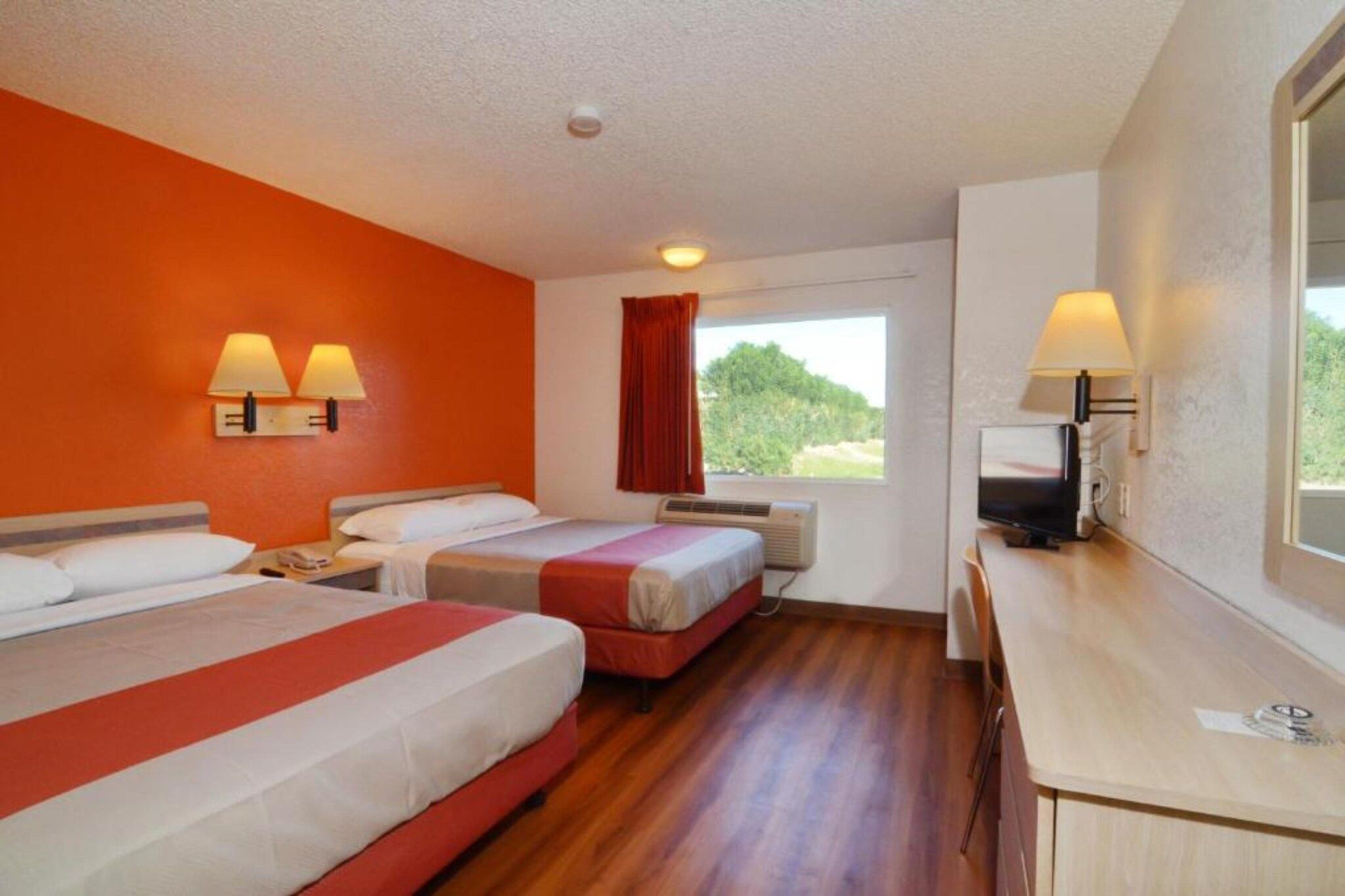 A hotel room with two beds, a desk, a TV, and a window with a view of greenery.