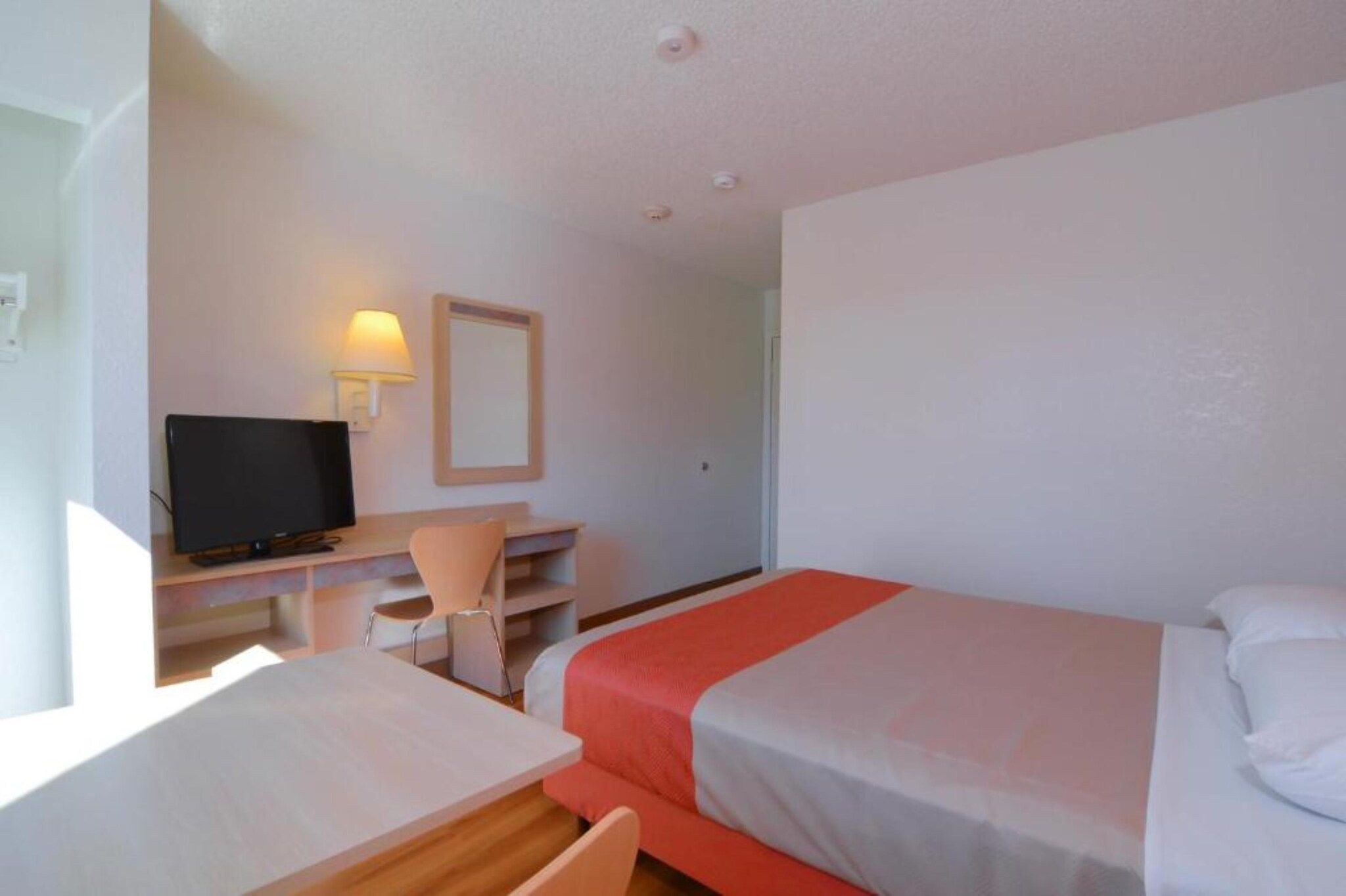Une chambre d’hôtel comprenant un lit, un bureau avec une chaise, une télévision et un lampadaire fixé au mur.