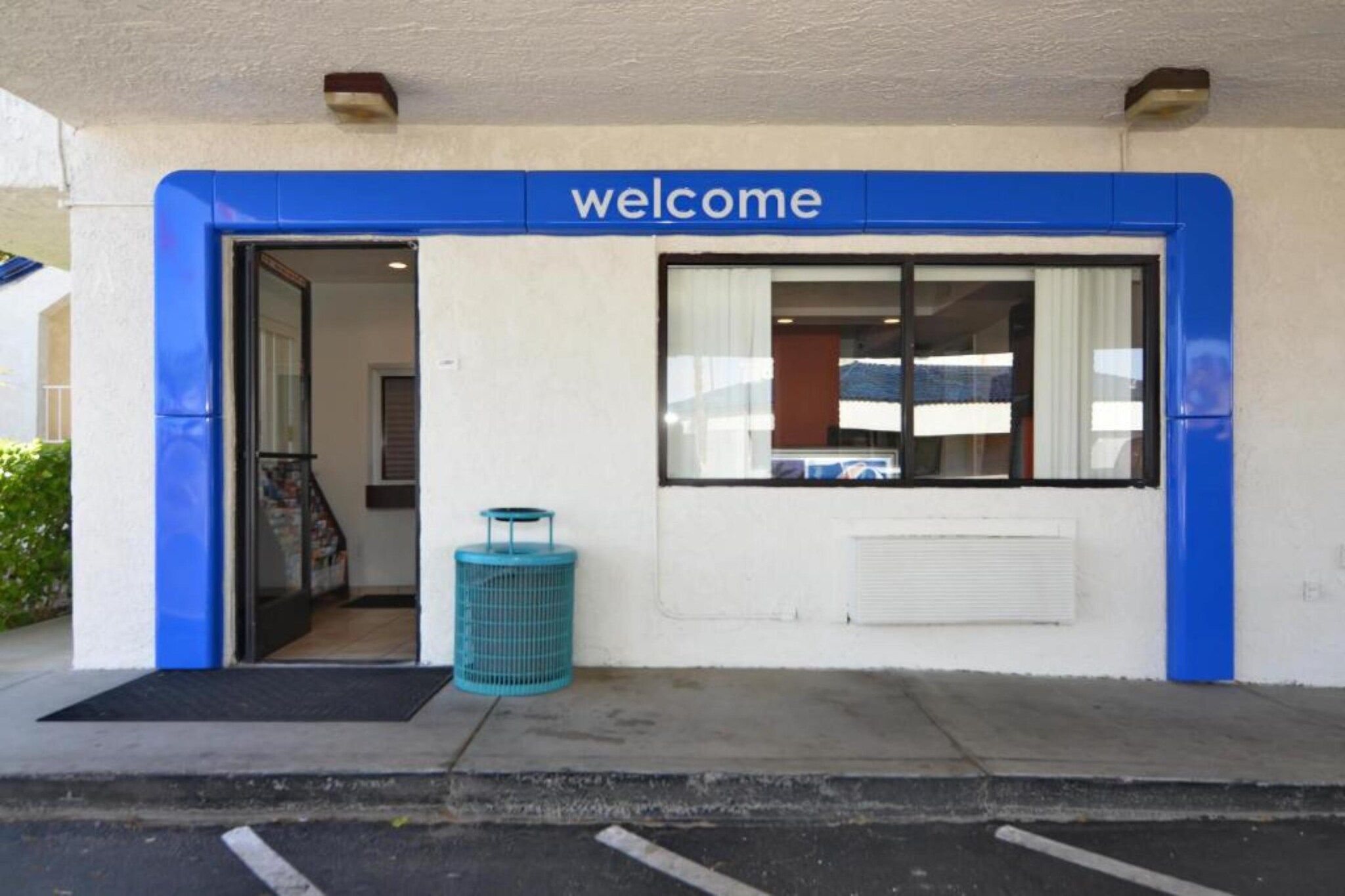 Une entrée d’immeuble avec une pancarte « Bienvenue », une porte en verre, une poubelle bleue et une fenêtre avec des stores.