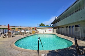 Piscina com escada de metal, cercada por uma grade, e um edifício com telhado azul ao fundo.