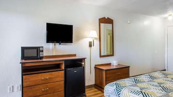 A hotel room with a bed, a desk with a microwave, a refrigerator, a television, a bedside table, and a mirror.