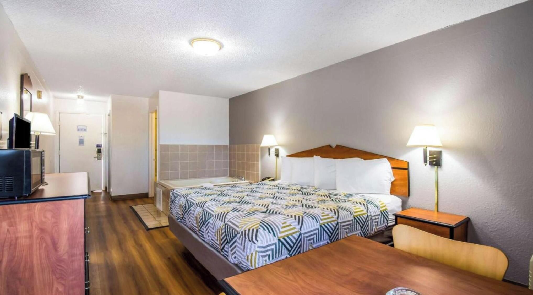 A hotel room with a bed, bedside table, lamp, and a desk with a television.