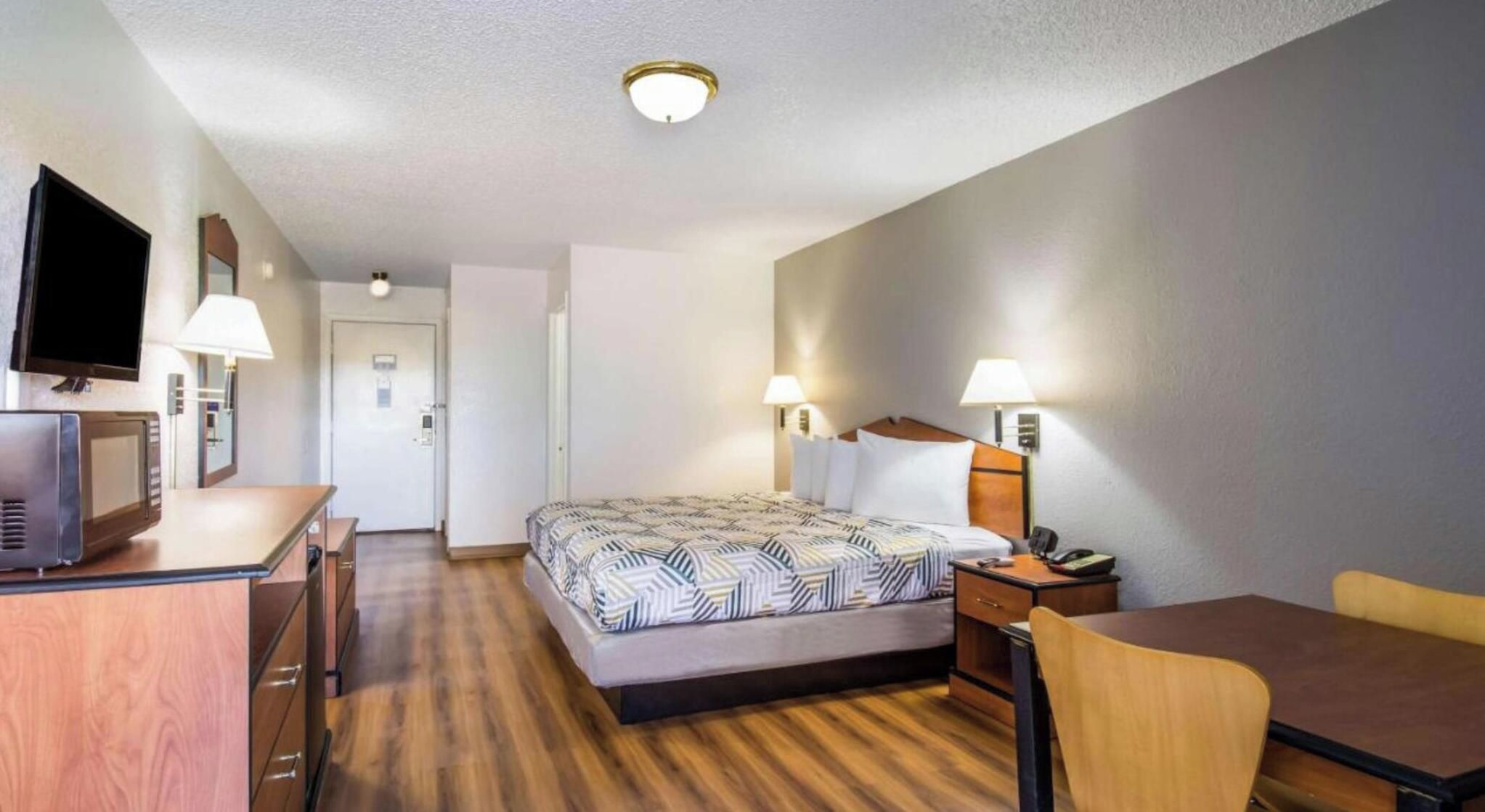Quarto de hotel com cama, escrivaninha, cadeira, televisão e luminárias.