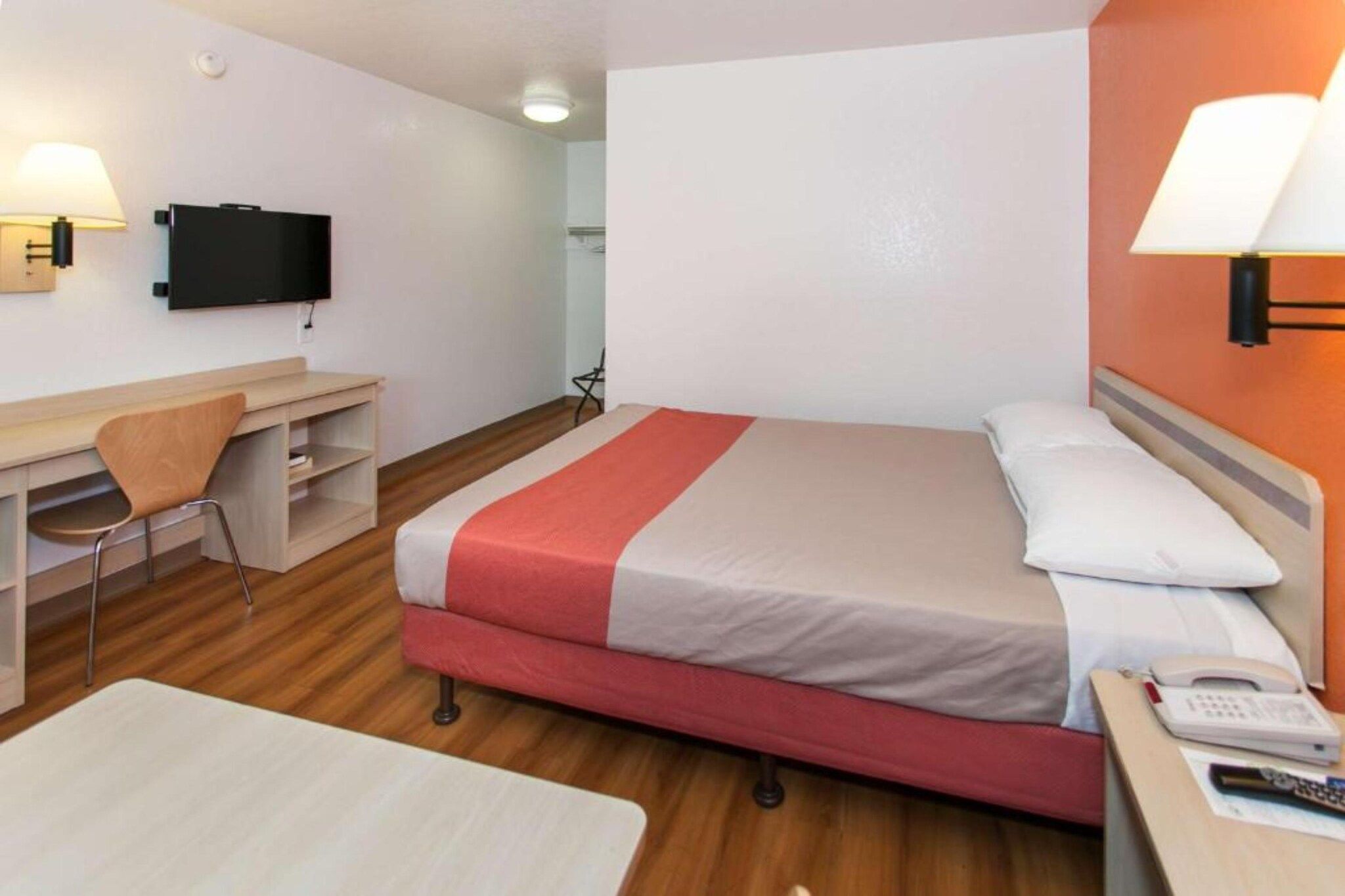A hotel room with a bed, a desk, a chair, a TV mounted on the wall, and a telephone on the bedside table.