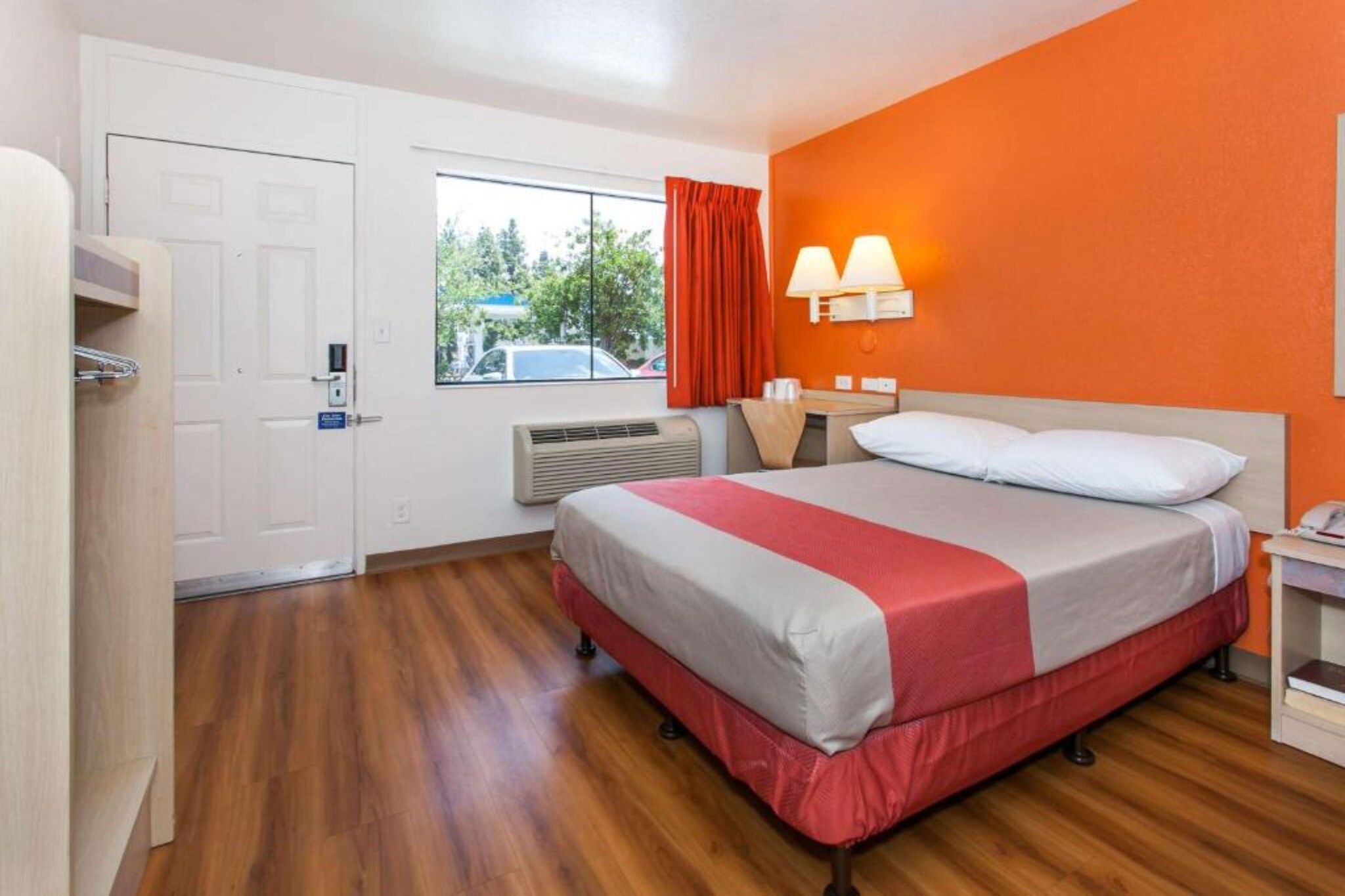 A hotel room with a bed, a window with curtains, an air conditioning unit, and a nightstand with a lamp.