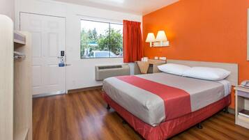 A hotel room with a bed, a window with curtains, an air conditioning unit, and a nightstand with a lamp.