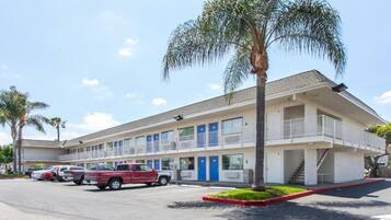 A motel with a two-story structure, a parking lot with several vehicles, and a palm tree in front.