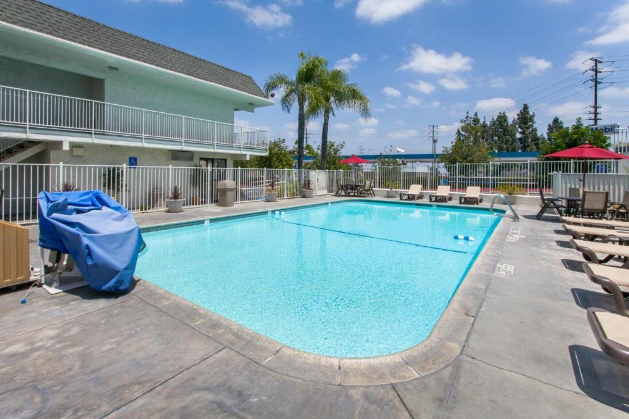 A swimming pool with lounge chairs and a covered area, surrounded by a fence and a building with a balcony.