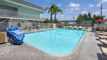 A swimming pool with lounge chairs and a covered area, surrounded by a fence and a building with a balcony.