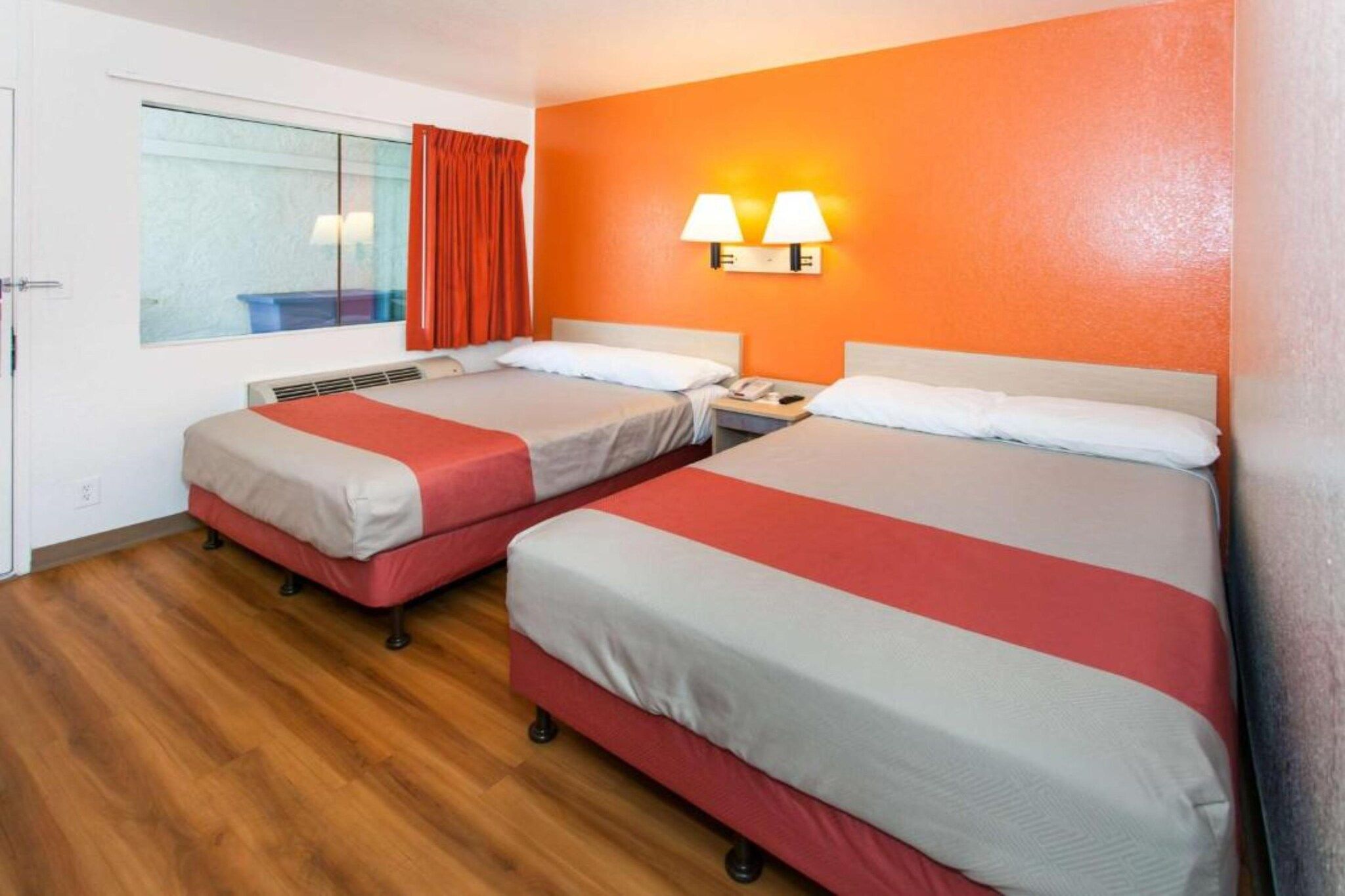 A hotel room with two beds, a window with curtains, a wall-mounted lamp, and a wooden floor.
