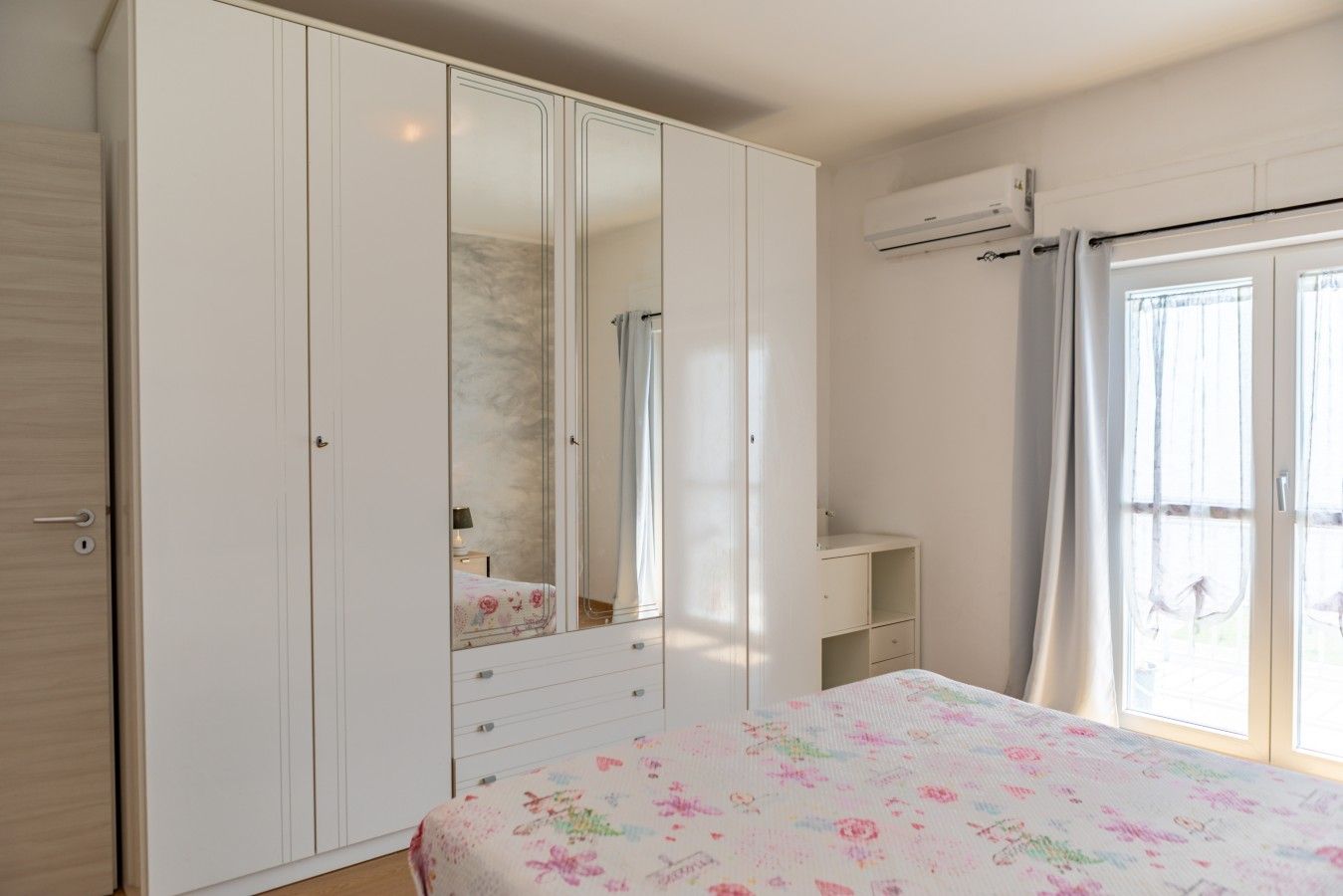 Een slaapkamer met een grote inbouwkast, een bed met bloemenprint beddengoed, een klein nachtkastje en een raam met gordijnen.