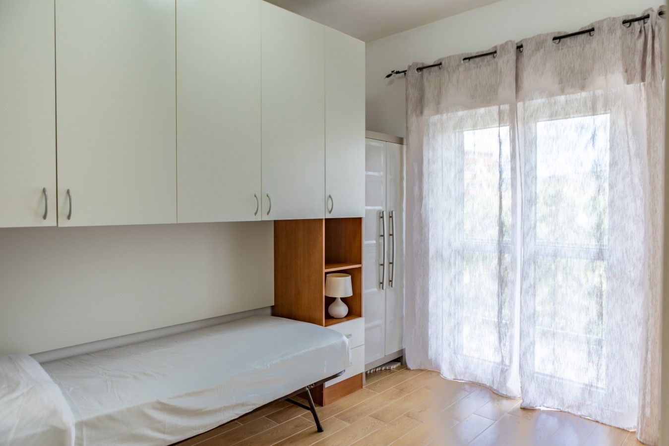 A room with a single bed, a wooden shelf with a lamp, white cabinets, and a window with sheer curtains.