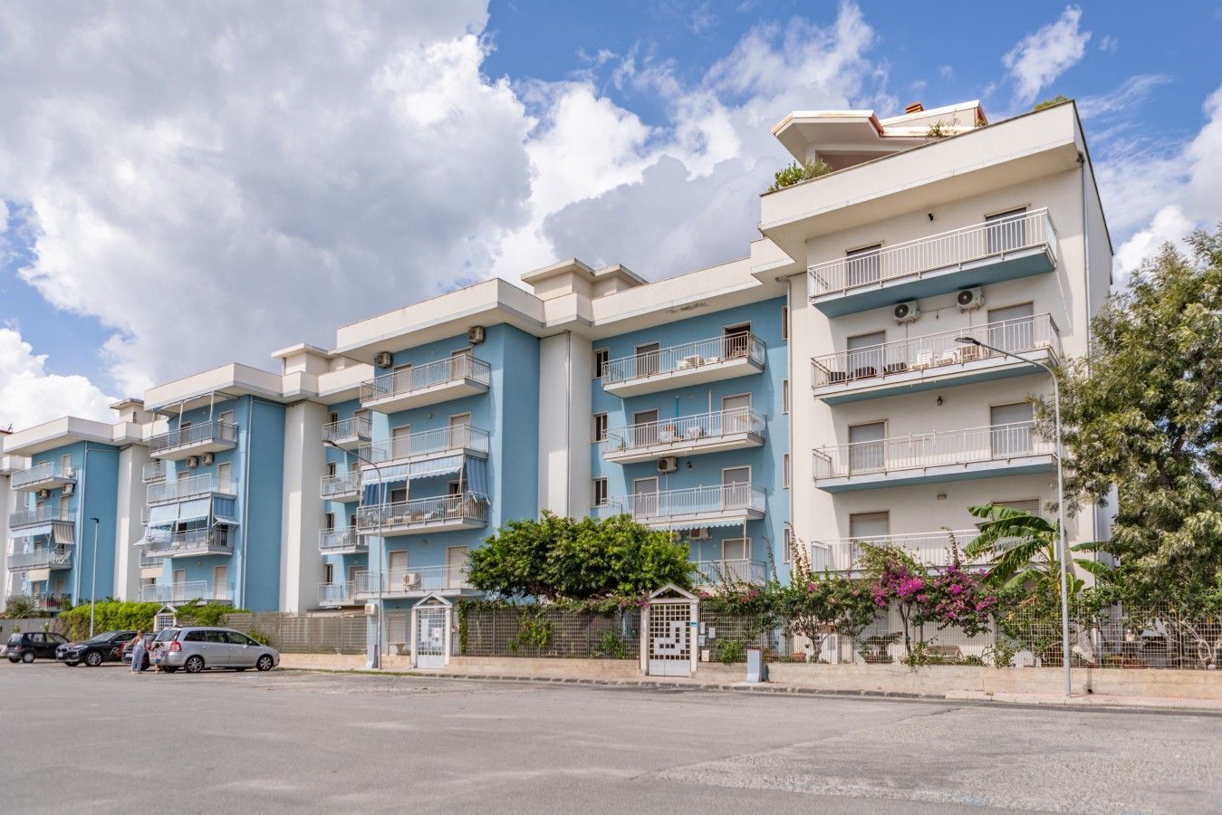A multi-story residential building with balconies, a parking area, and a clear sky.