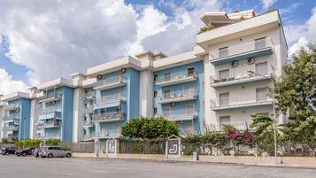 A multi-story residential building with balconies, a parking area, and a clear sky.