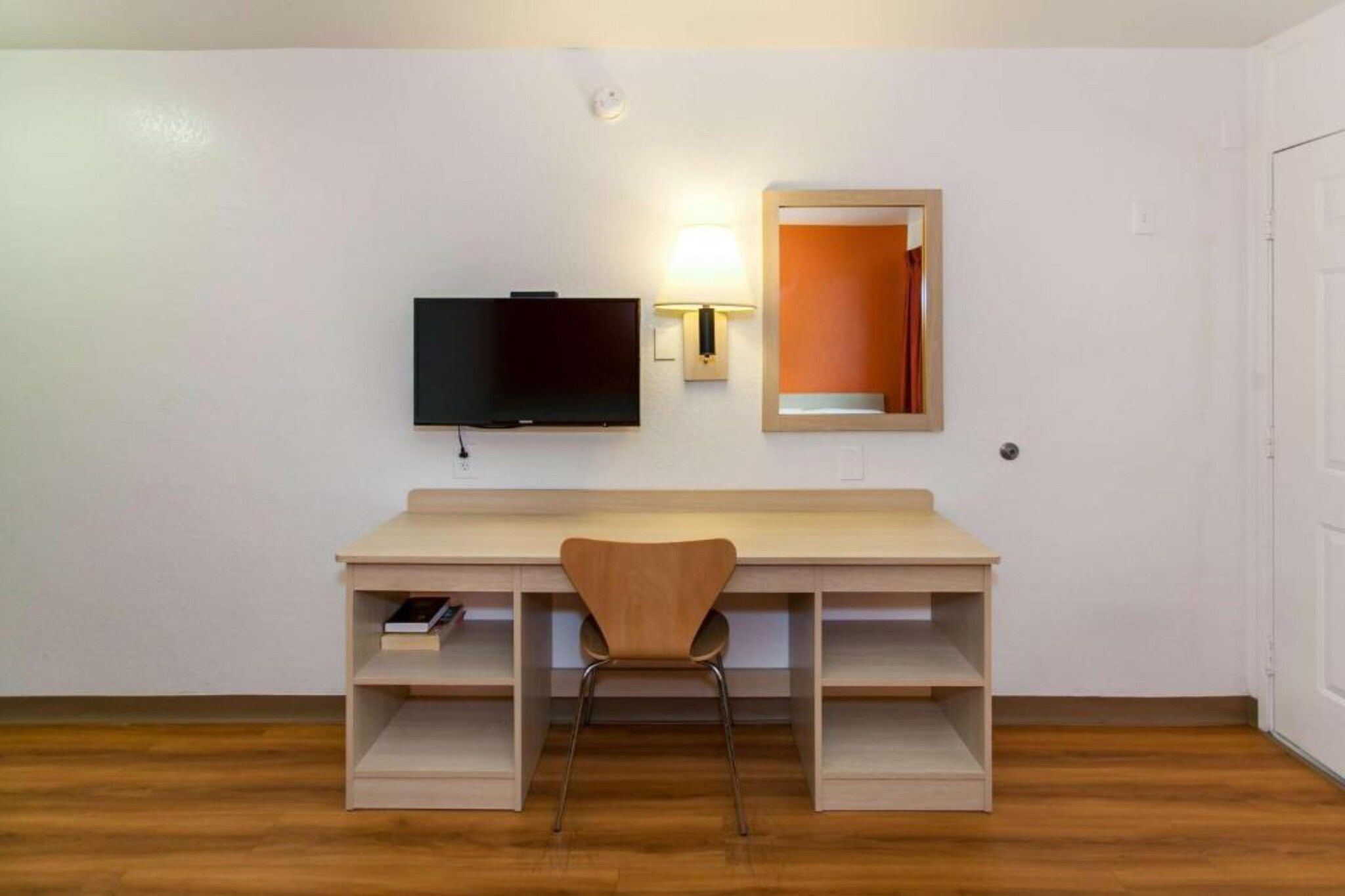 A room with a wooden desk, a chair, a television mounted on the wall, and a mirror.