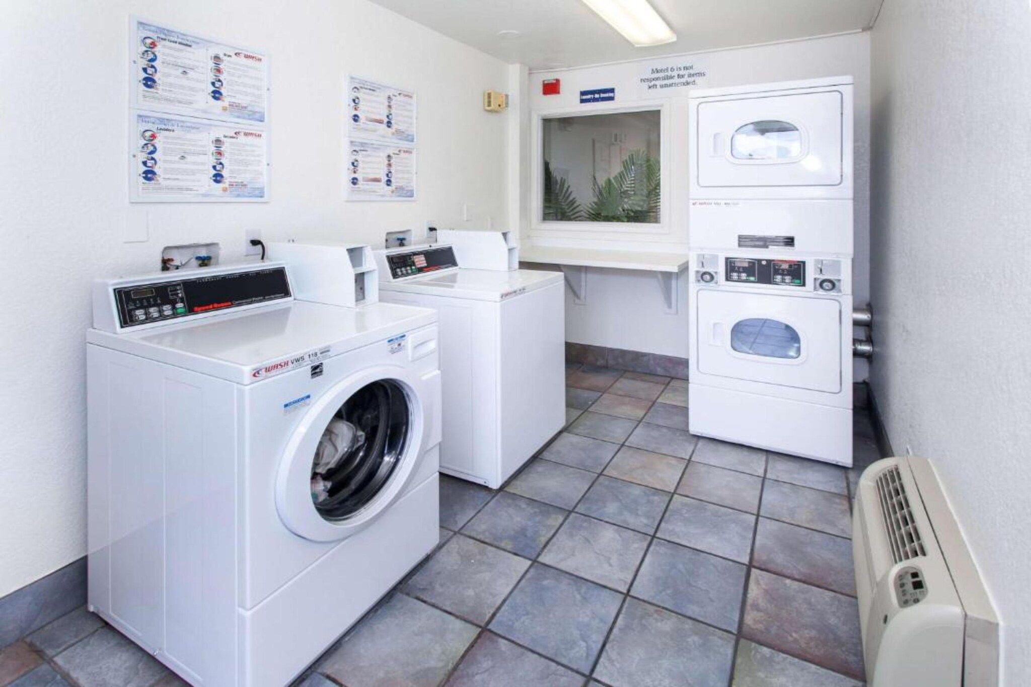 A laundry facility with a front-loading washer and dryer, a small window with a plant, and informational posters on the wall.