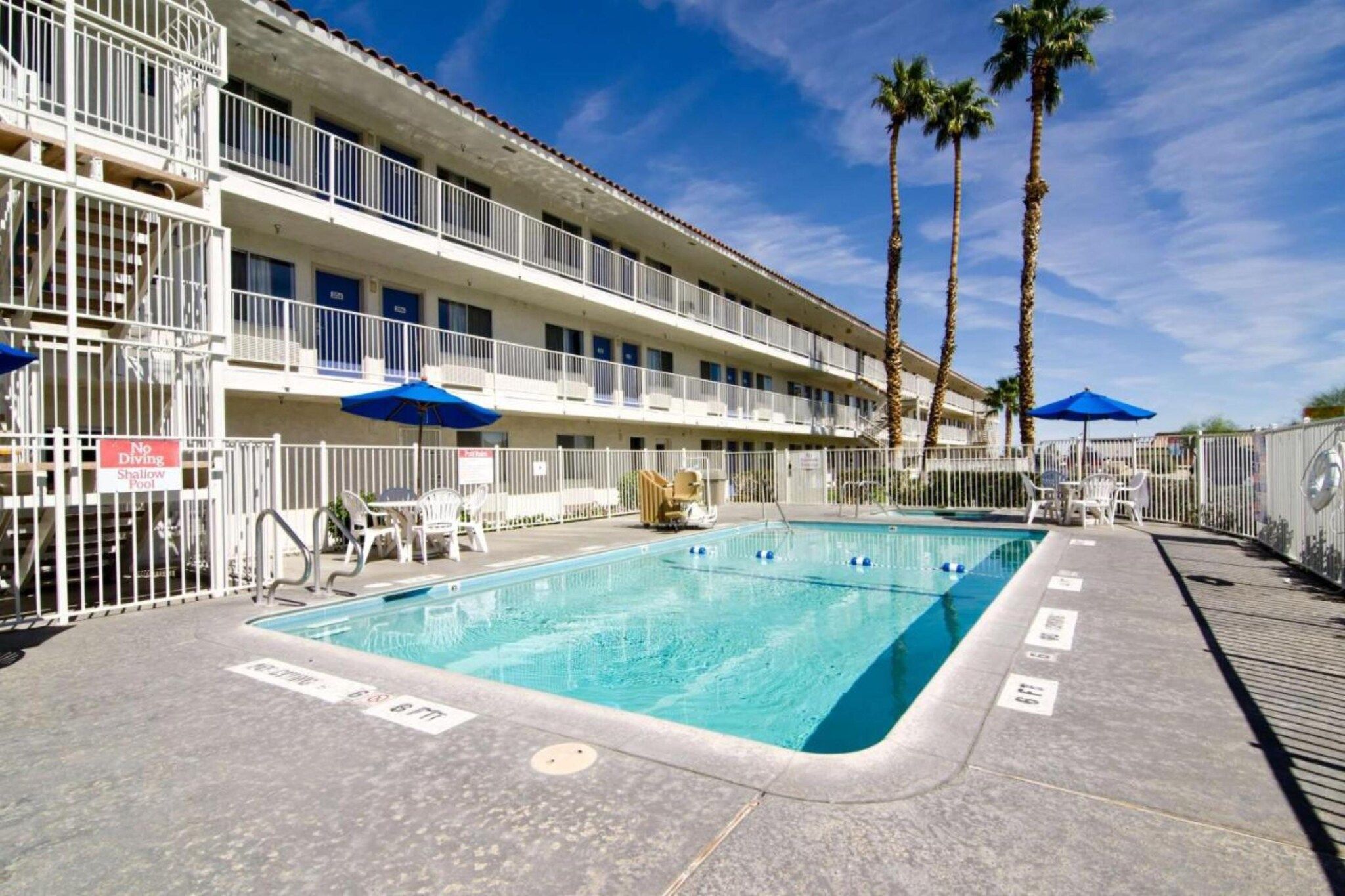 A hotel pool area with a clear blue pool, white poolside chairs, and palm trees.