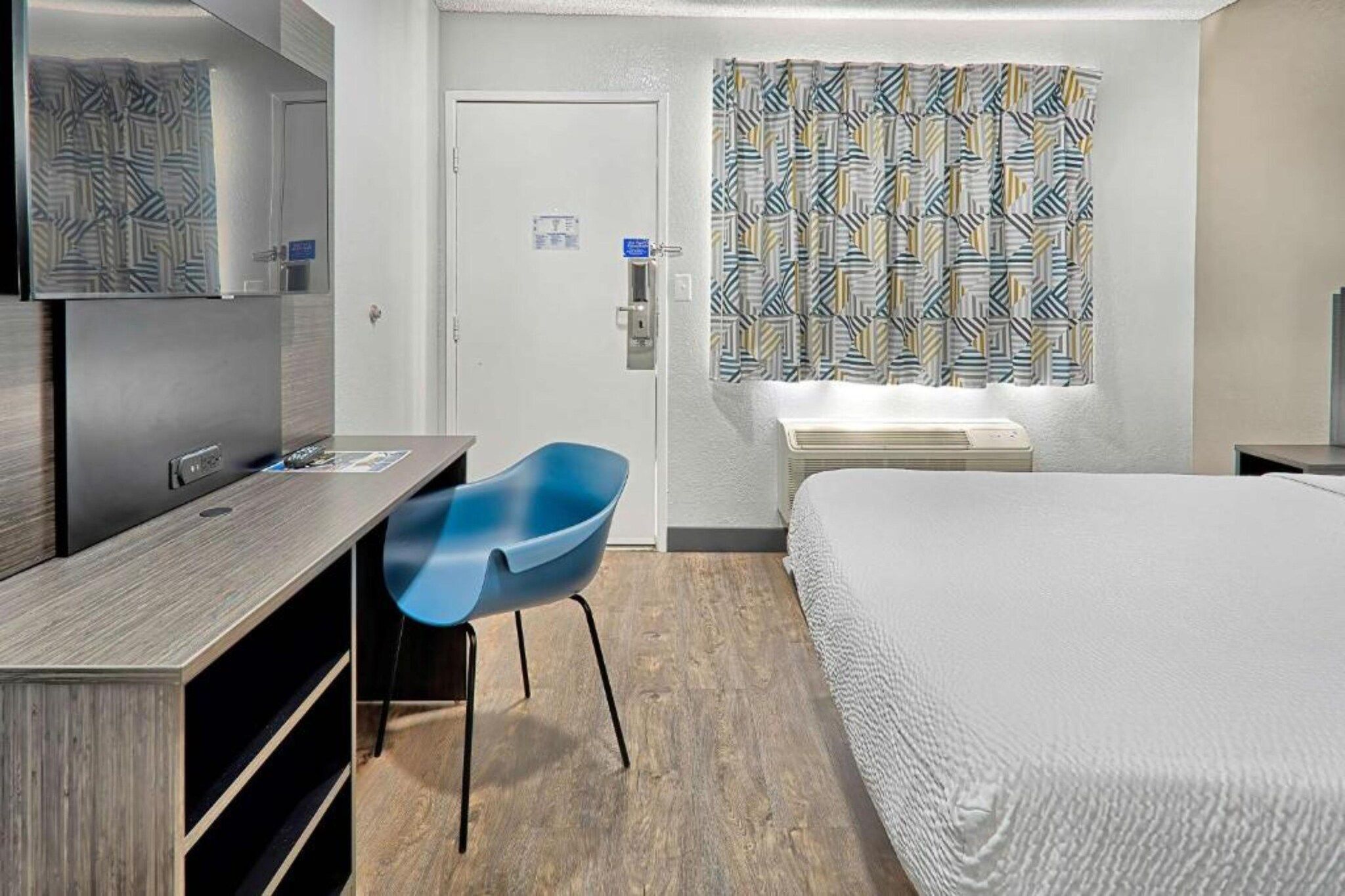 A hotel room with a bed, a desk, a chair, a window with patterned curtains, and a wall-mounted air conditioning unit.