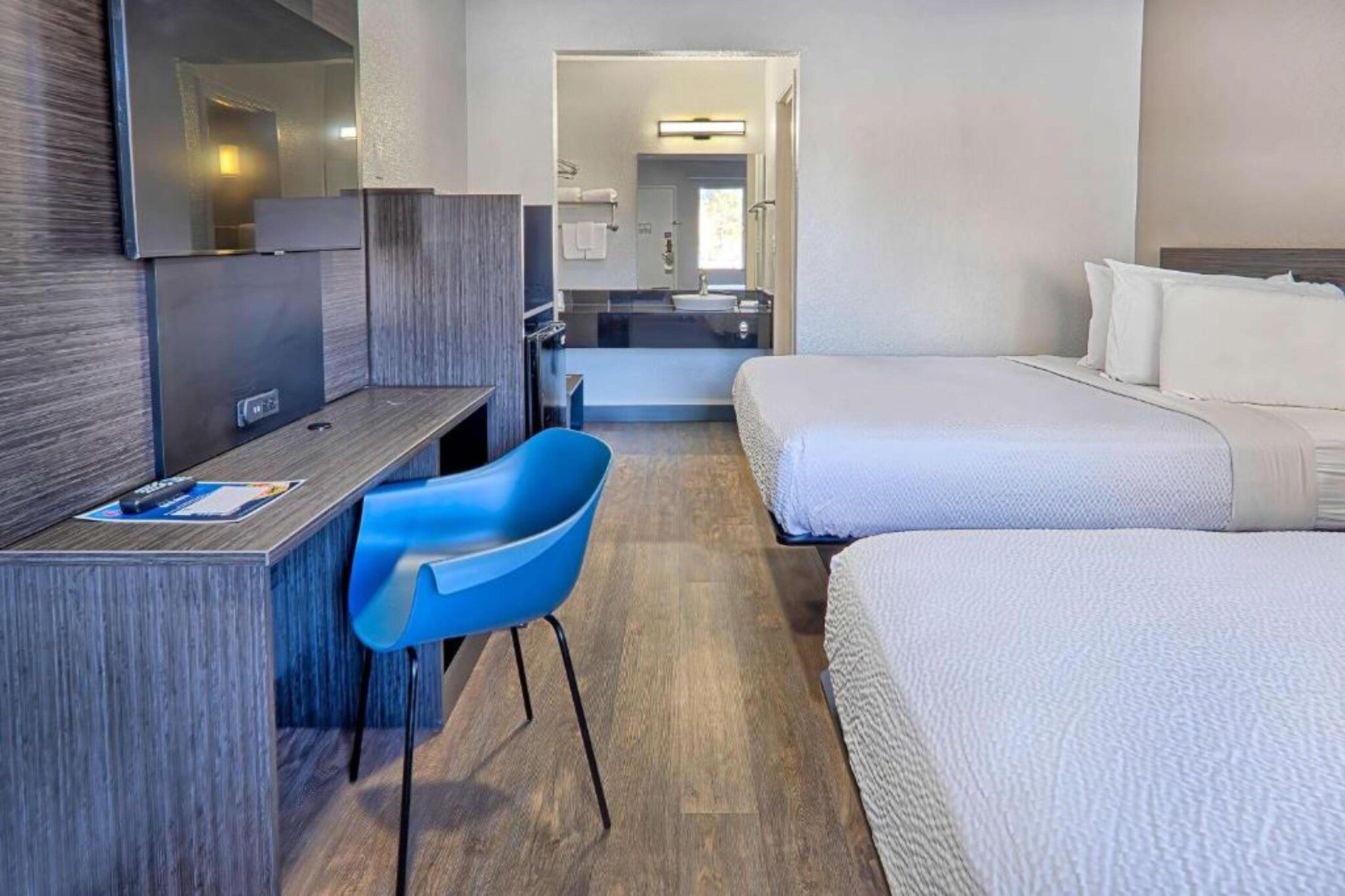 Een hotelkamer met een bureau, een blauwe stoel, twee bedden en een badkamer die door een open deur zichtbaar is.
