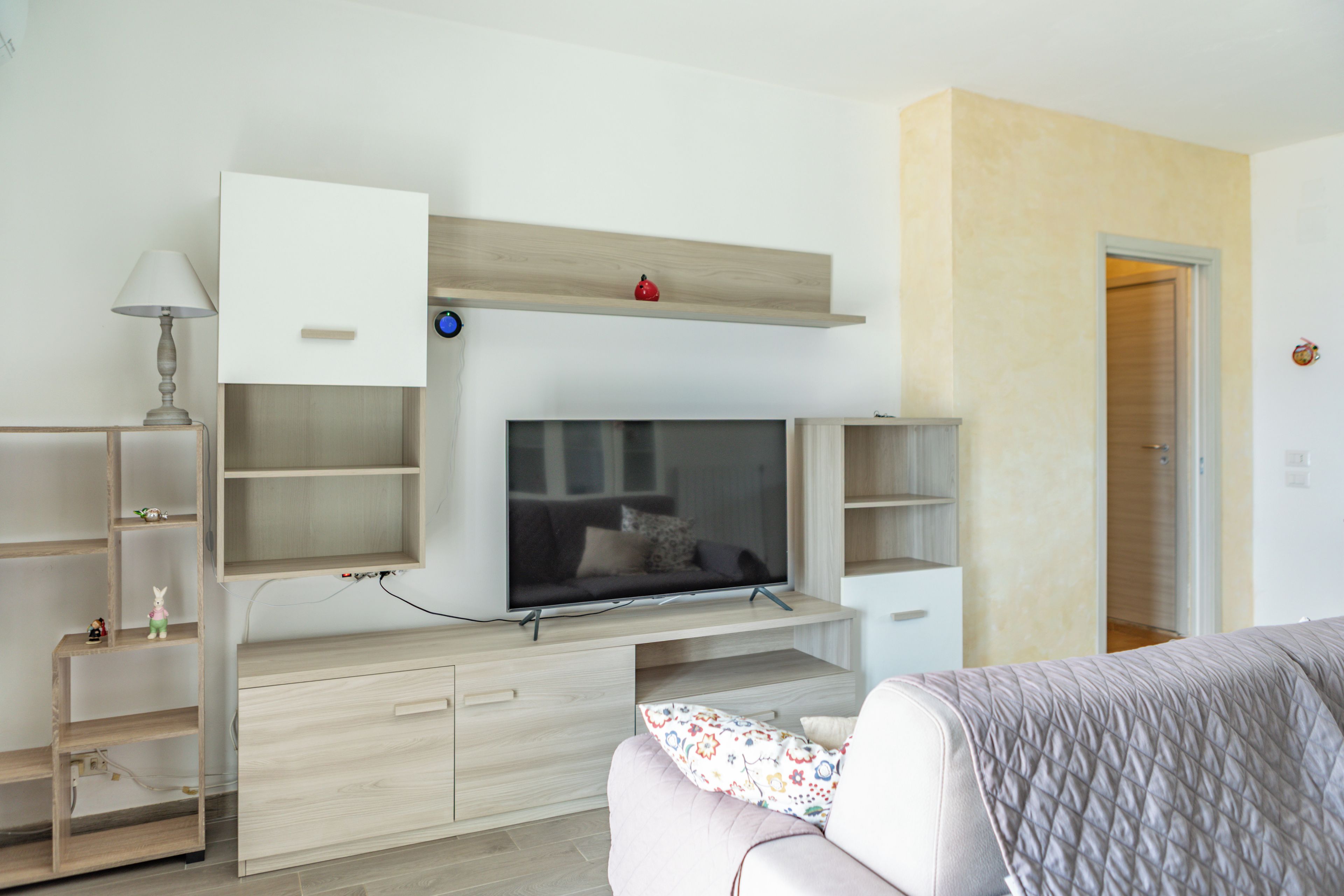 Ein modernes Schlafzimmer mit Fernseher an einer Holzeinrichtung, einem Nachttisch mit Lampe und einem Bett mit gemustertem Überwurf.