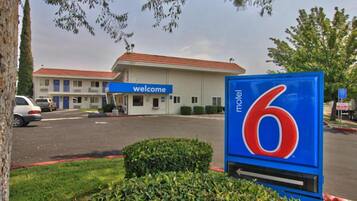 A motel with a 'Welcome' sign, a '6' sign indicating the number of rooms, and a parking lot with cars.
