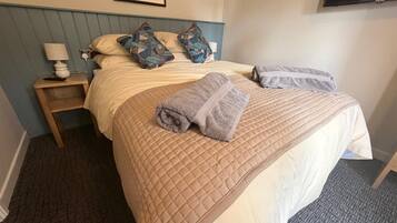 Una habitación de hotel con cama, almohadas, una mesita de noche, una lámpara y un cuadro en la pared.