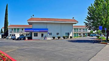 Un motel de dos pisos con techo de tejas rojas, un letrero azul que dice 'Welcome' y un estacionamiento con varios autos.