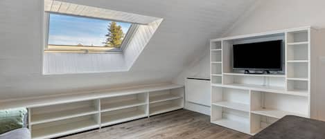 Una habitación con techo inclinado, tragaluz, televisor de pantalla plana sobre un estante blanco y sofá gris.