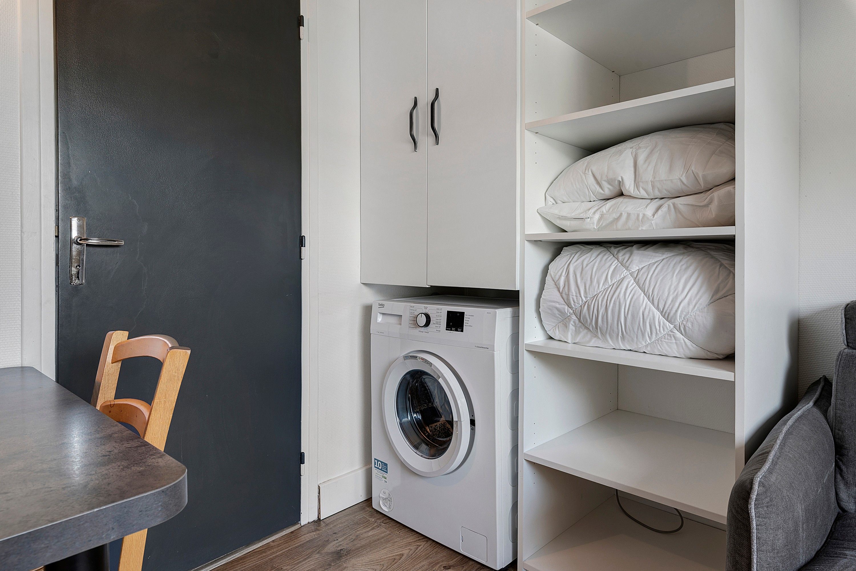 Un lavadero pequeño con lavadora, una silla y estantería con ropa de cama doblada.