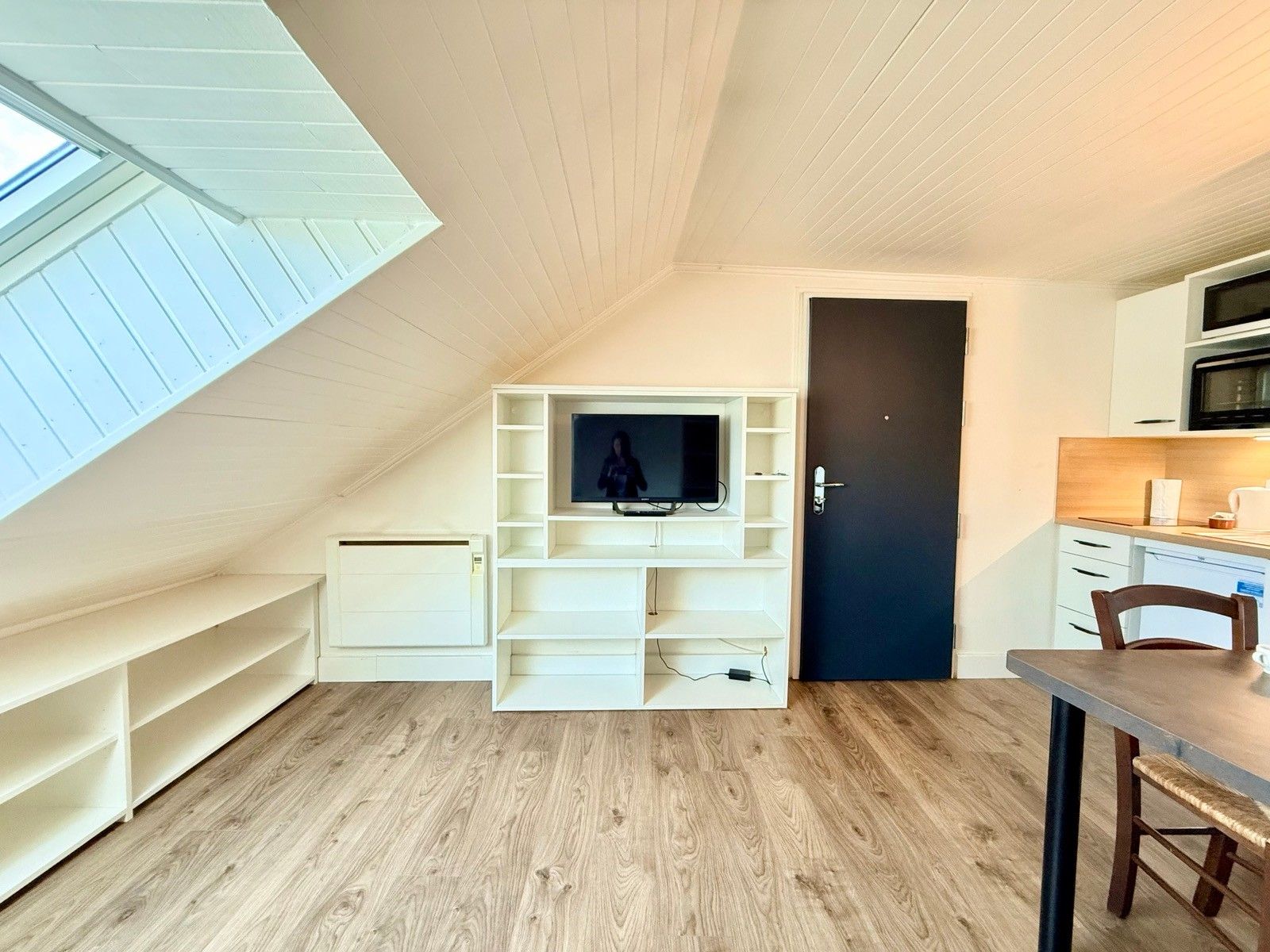 Een compacte kamer met schuine kap, een inbouwkast voor de televisie, een donkere deur en een kitchenette.