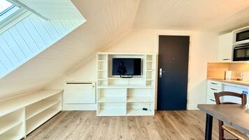 Een compacte kamer met schuine kap, een inbouwkast voor de televisie, een donkere deur en een kitchenette.