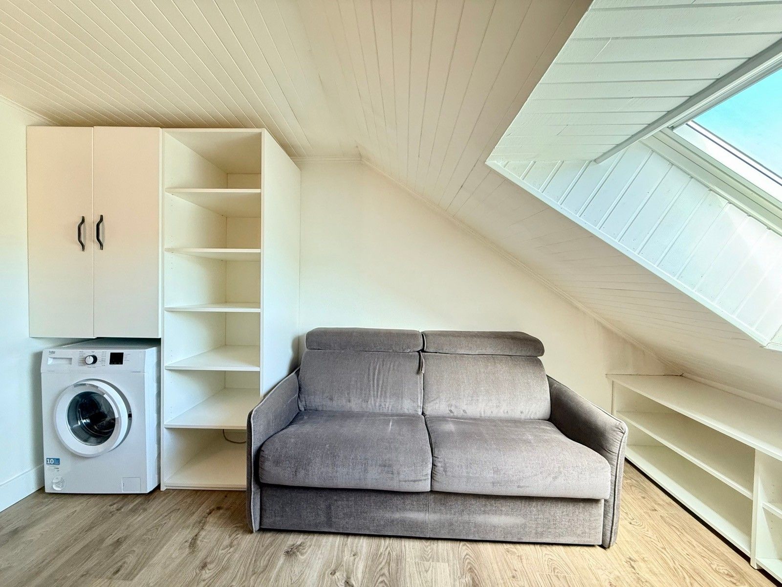 Ein kompakter Wohnraum mit grauem Sofa, Waschmaschine und einer eingebauten Regallösung.