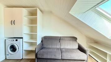 Ein kompakter Wohnraum mit grauem Sofa, Waschmaschine und einer eingebauten Regallösung.