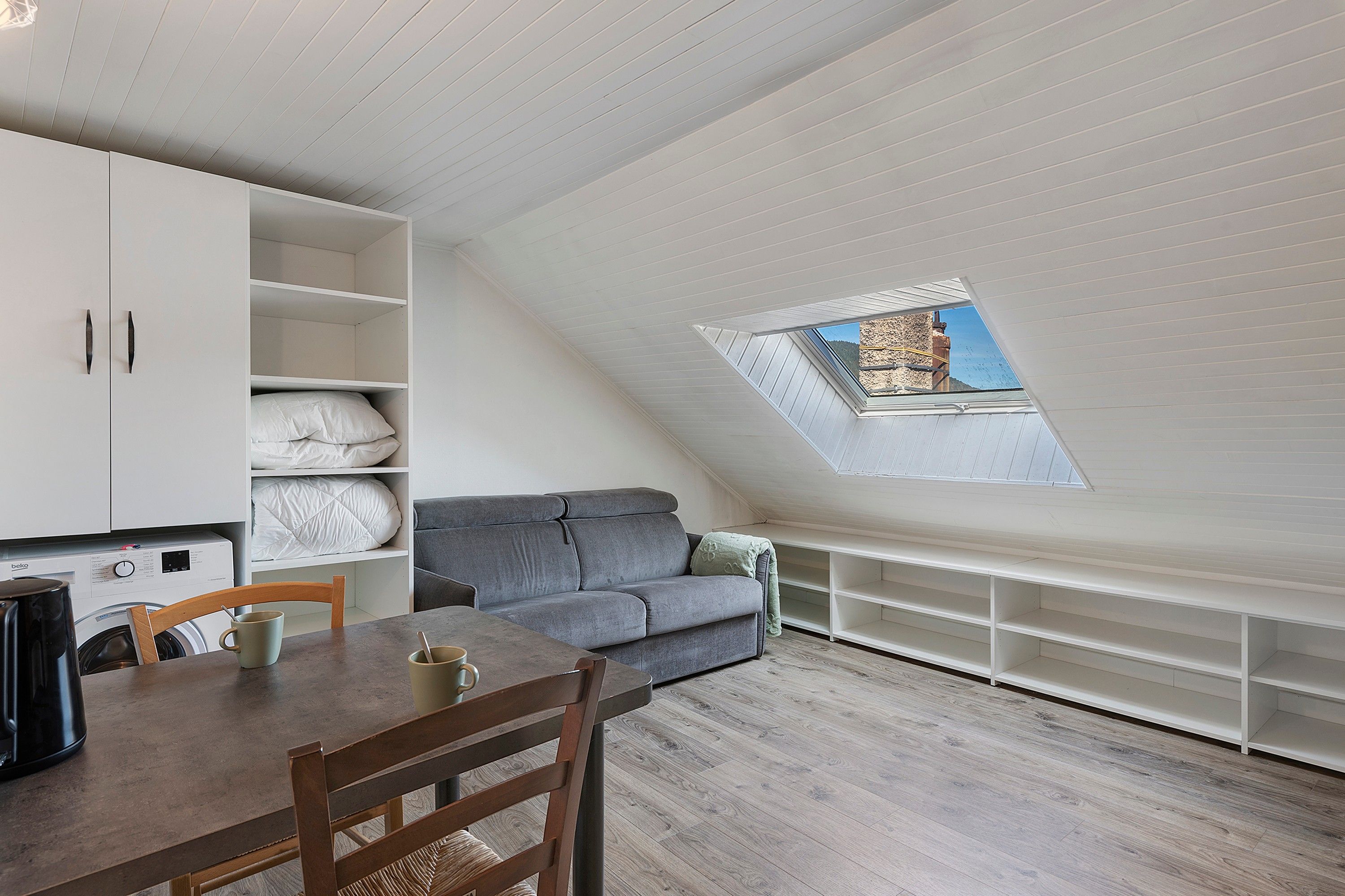 Un espace de vie compact doté d’un plafond en pente, d’un canapé intégré, d’une table à manger avec des chaises et d’un petit coin cuisine.
