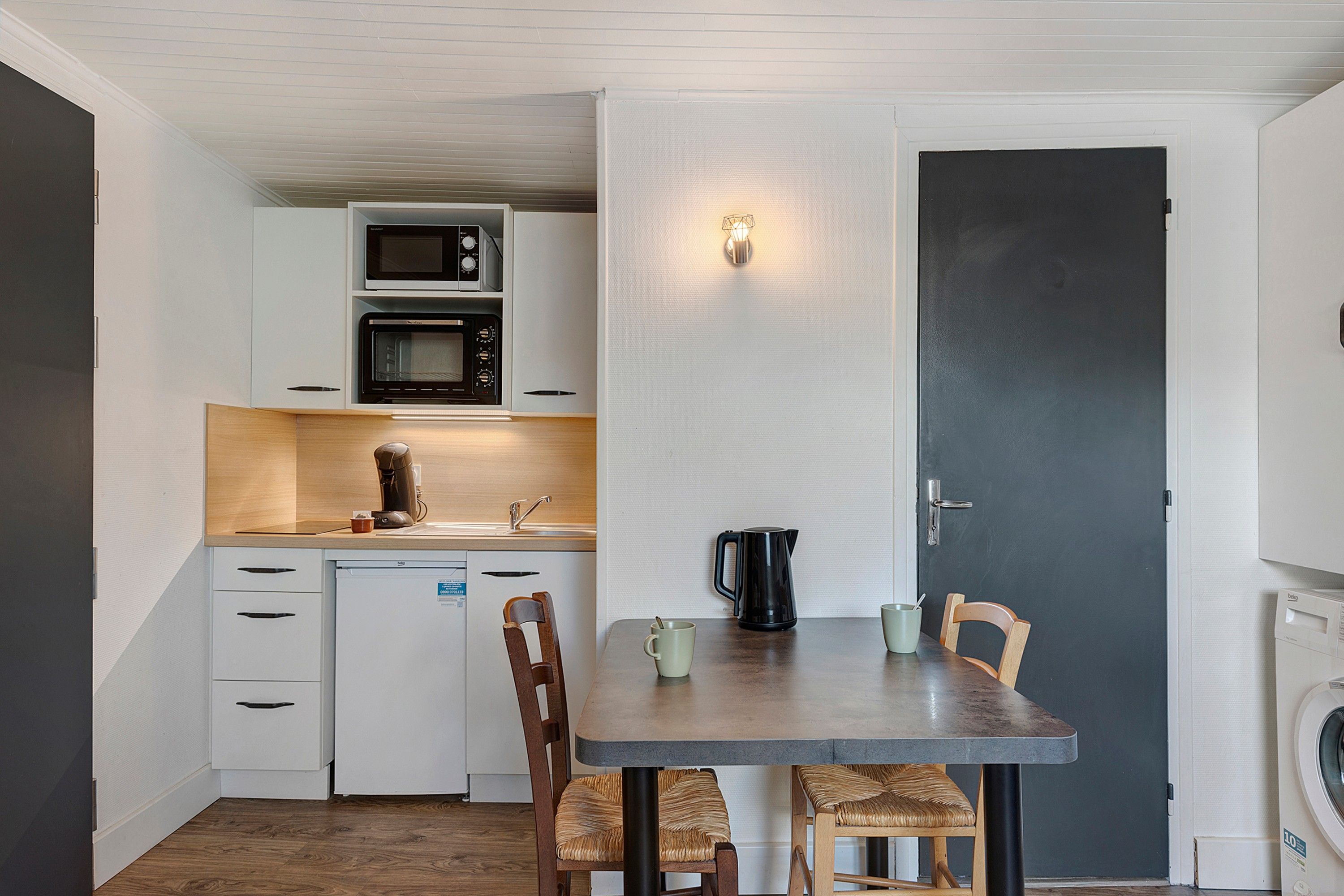 A compact kitchen with a microwave, oven, and coffee machine. There is a dining table with chairs and a kettle.
