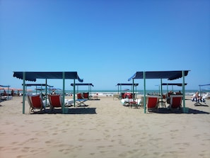 Ein Strand mit zwei überdachten Sitzbereichen, jeweils mit mehreren Stühlen und Tischen.