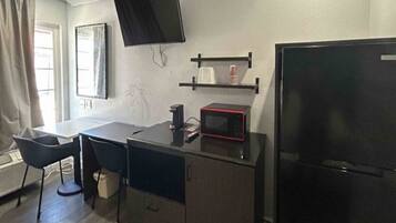 A compact kitchenette with a microwave, a small refrigerator, a flat-screen TV mounted on the wall, and a window with curtains.