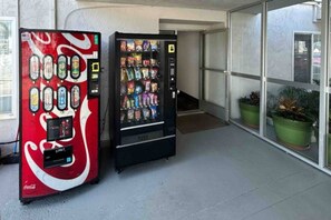 Dos máquinas expendedoras, una con el logotipo de Coca-Cola y la otra con diferentes bocadillos, ubicadas fuera de un edificio con puerta de cristal y plantas en macetas.