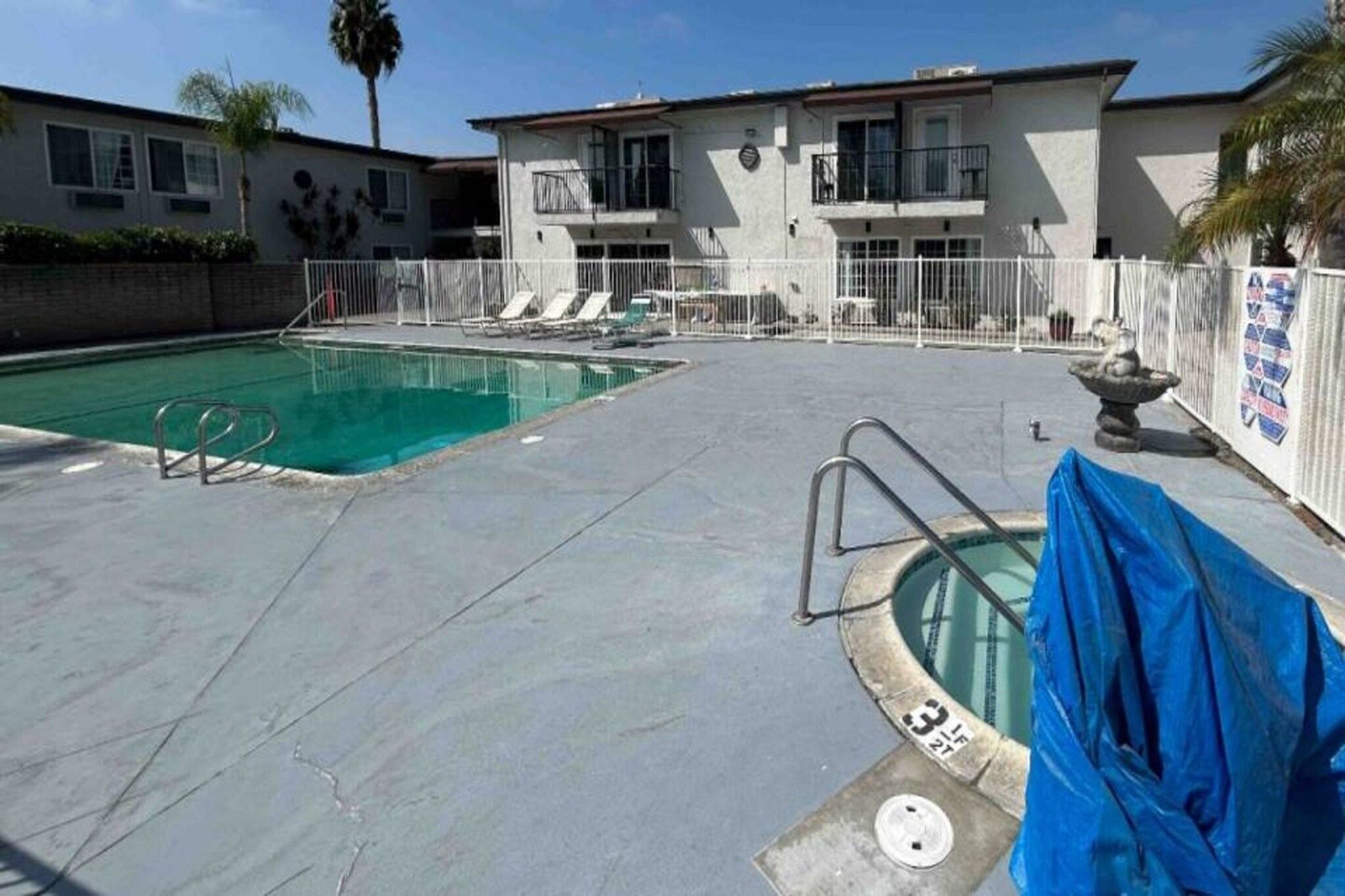 A swimming pool with a covered area, a fenced deck, and a building with multiple windows and balconies.