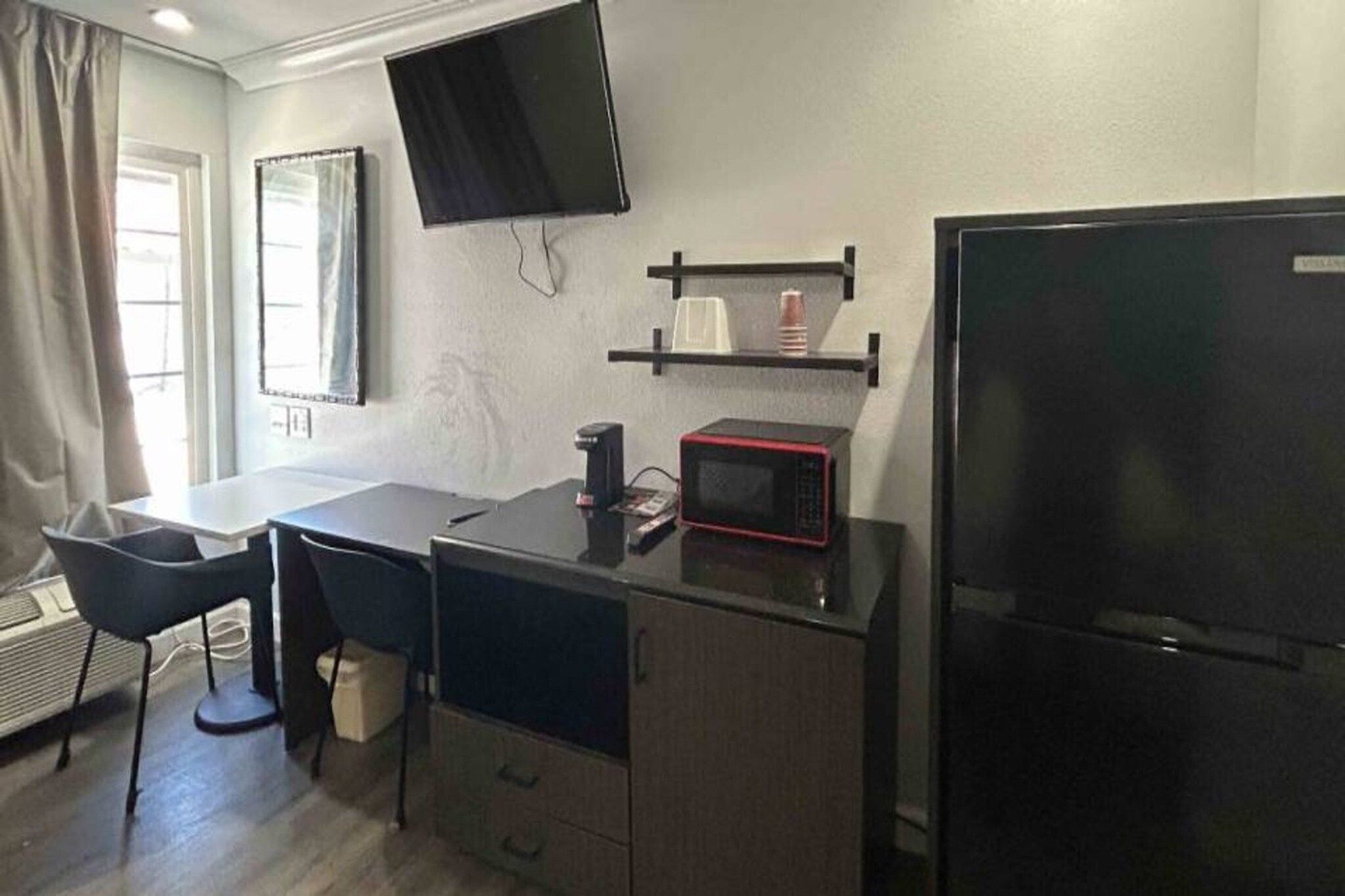 A compact kitchenette with a microwave, a small refrigerator, a flat-screen TV mounted on the wall, and a window with curtains.