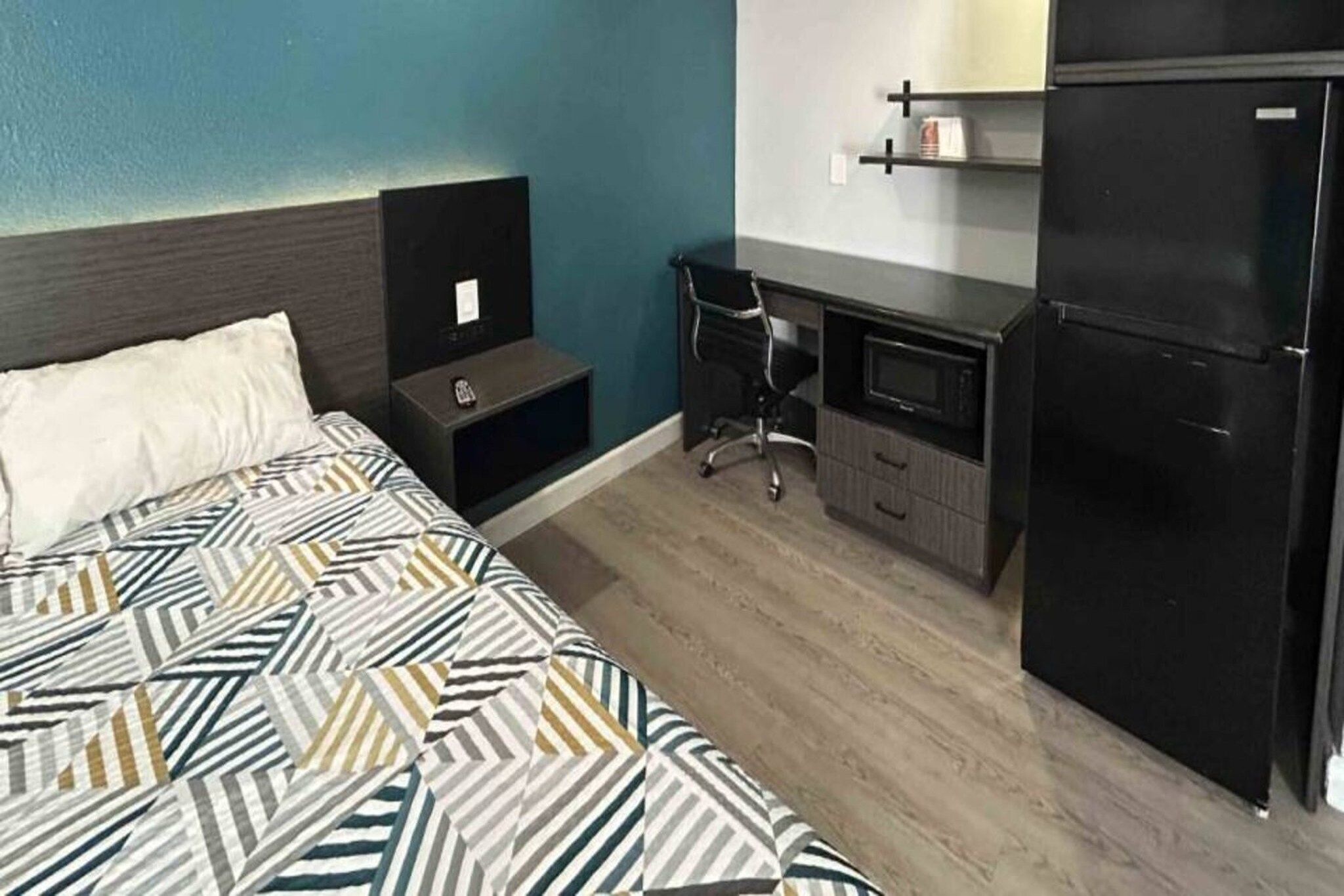A hotel room with a bed, a desk, a chair, a refrigerator, and a nightstand.